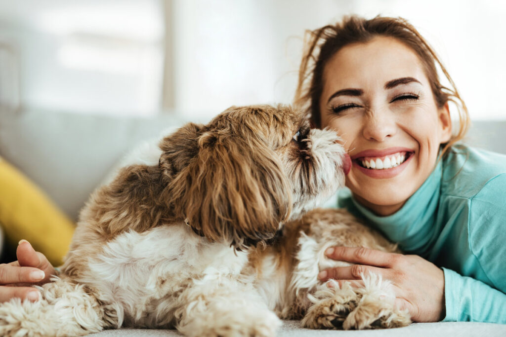Young woman having fun while her dog is kissing her at home.